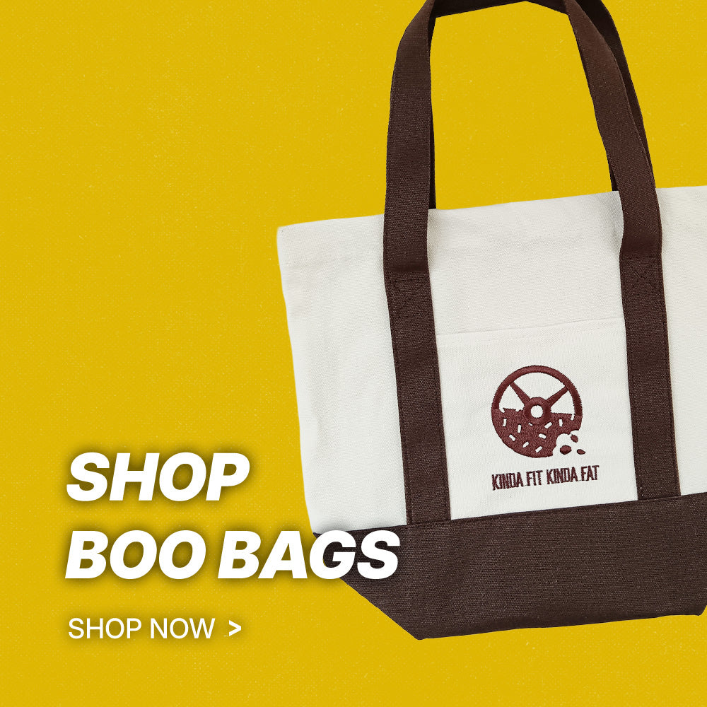 A white canvas tote bag with dark brown handles and bottom trim, featuring a brown logo that reads 'KINDA FIT KINDA FAT' on the front pocket. The bag is set against a solid yellow background with promotional text on the left reading 'SHOP BOO BAGS' and 'SHOP NOW >'.